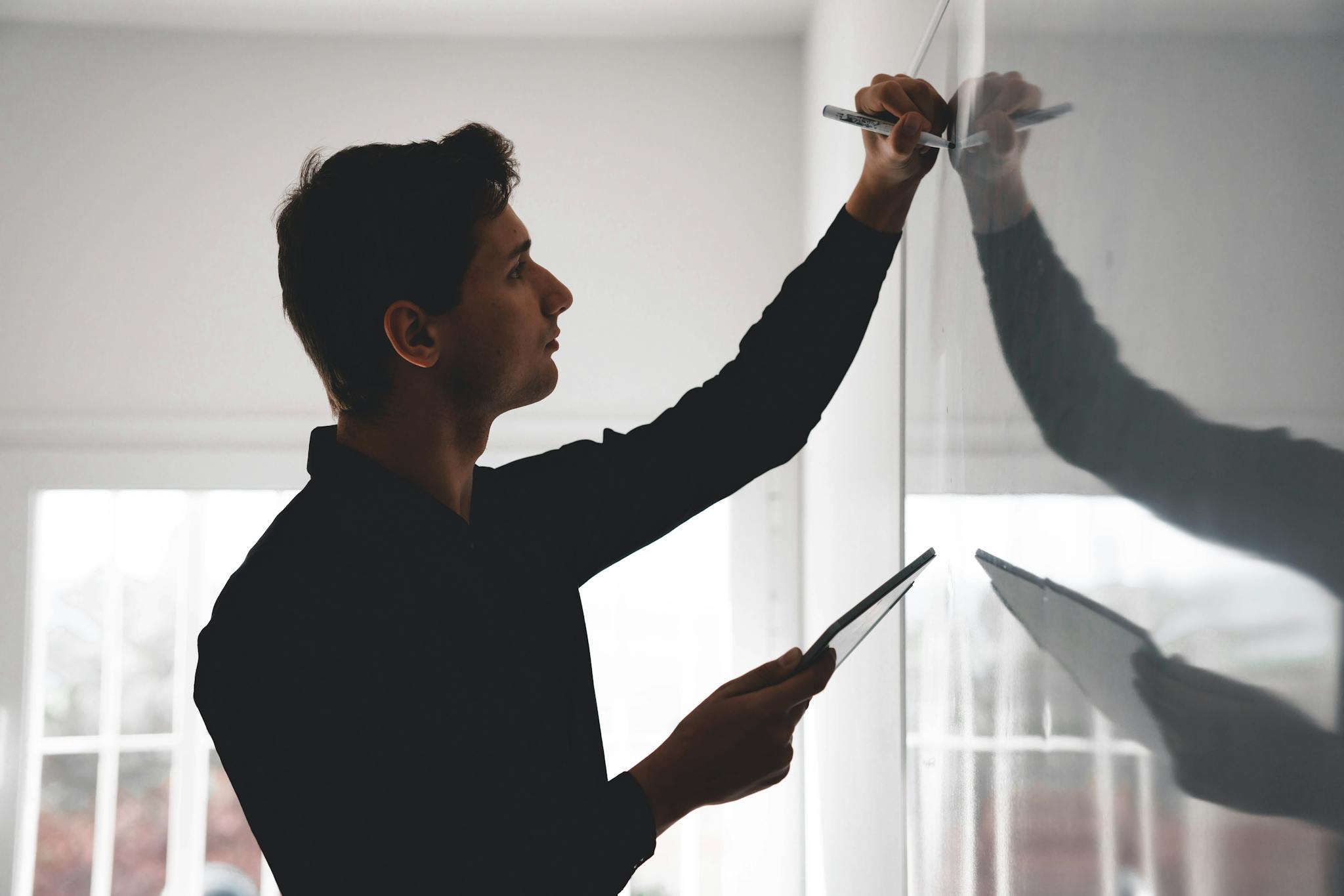 A man writing on a whiteboard while holding a tablet in a well-lit room.